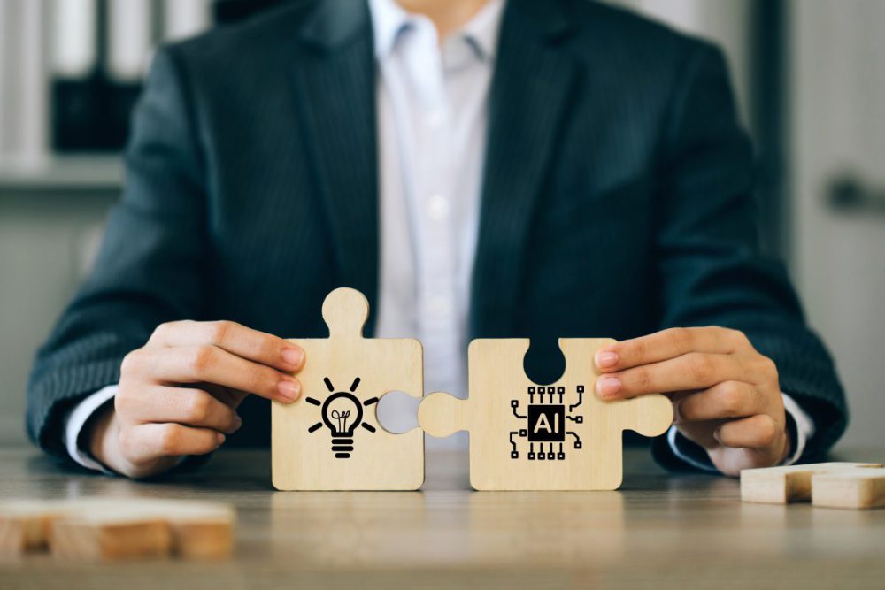 Das Ziel ist kein abruptes Umdenken, sondern die evolutionäre Weiterentwicklung bewährter Strategien. Person in Anzug hält zwei Puzzleteile aus Holz: auf dem einen ist ein Icon für eine Glühbirne, auf dem anderen eins für AI zu sehen.